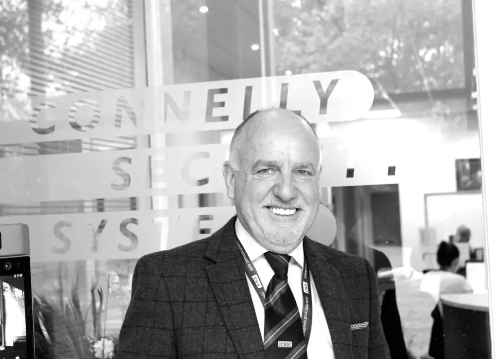 Black and white photo of Paul Connelly smiling and standing in front of a glass door that says Connelly Security Systems.