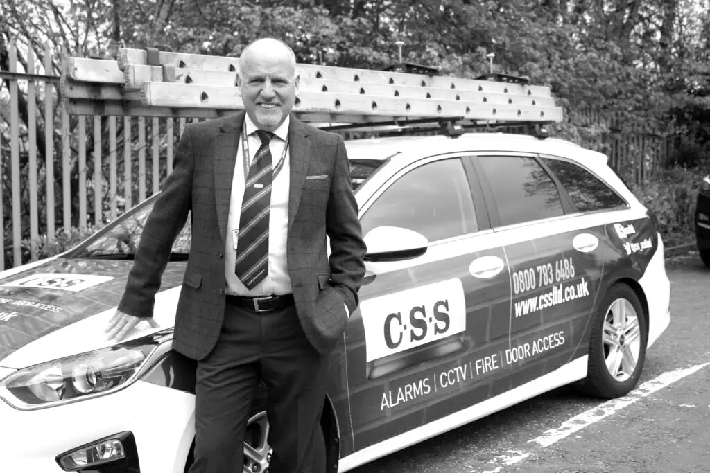 Black and white photo of Paul Connelly standing smiling and resting his right hand on the bonnet of a CSS branded car.
