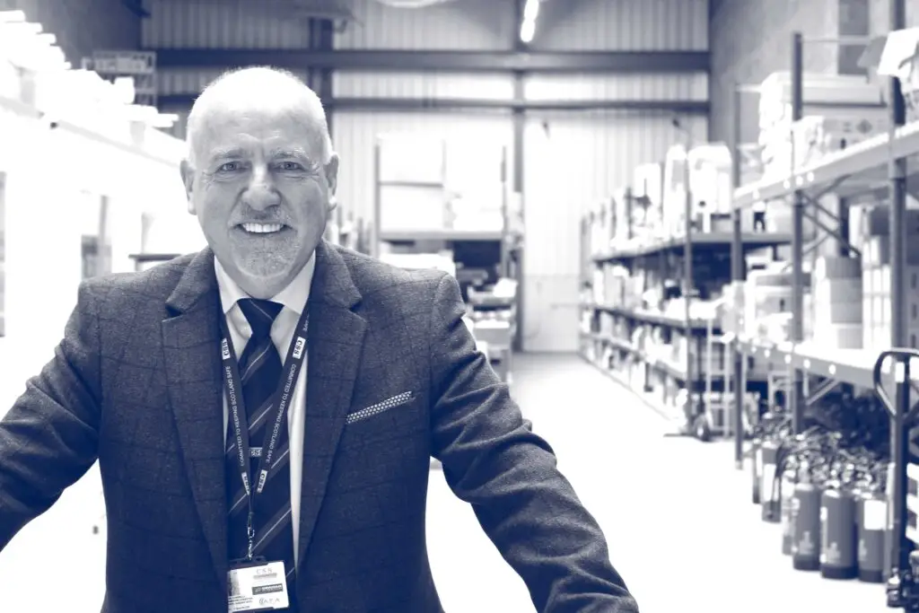Black and white photo of Paul Connelly smiling and standing in a warehouse. Shelves, boxes and fire extinguishers are visible behind him.