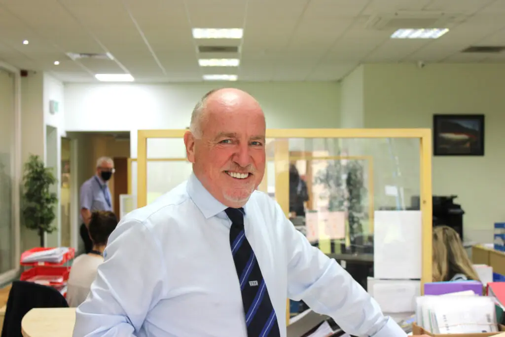 Photo of Paul Connelly standing in CSS office resting his left hand against a desk. Members of staff working to social distance measures can be seen behind him, one of whom is wearing a black face mask. There are safety separator screens mounted to desks.