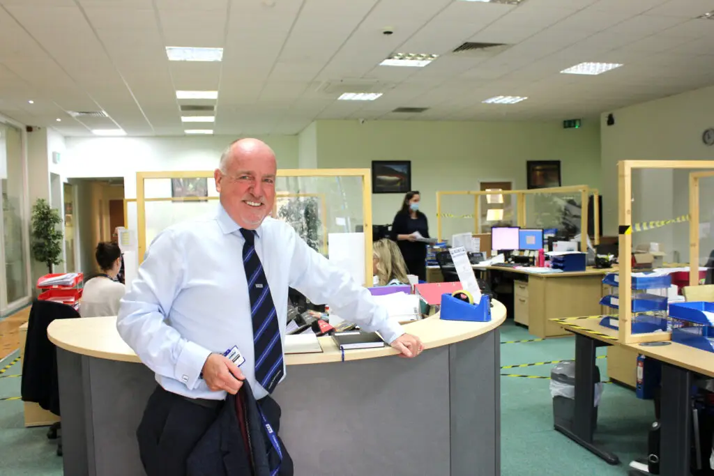 Photo of Paul Connelly standing in CSS office resting his left hand against a desk. Members of staff working to social distance measures can be seen behind him, one of whom is wearing a blue medical face mask. There are safety separator screens mounted to desks.