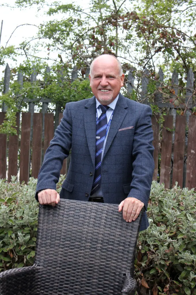 Photo of Paul Connelly smiling in garden location and resting his hands on the back of a chair.