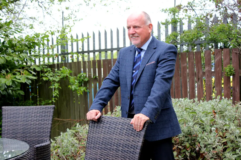 Photo of Paul Connelly smiling in garden location and resting his hands on the back of a chair.