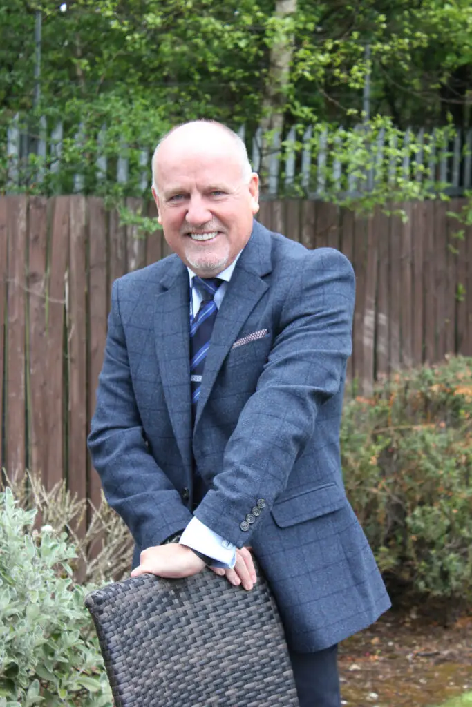 Photo of Paul Connelly smiling in garden location and resting his hands on the back of a chair.