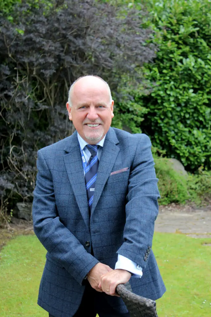 Photo of Paul Connelly smiling in garden location and resting his hands on the back of a chair.