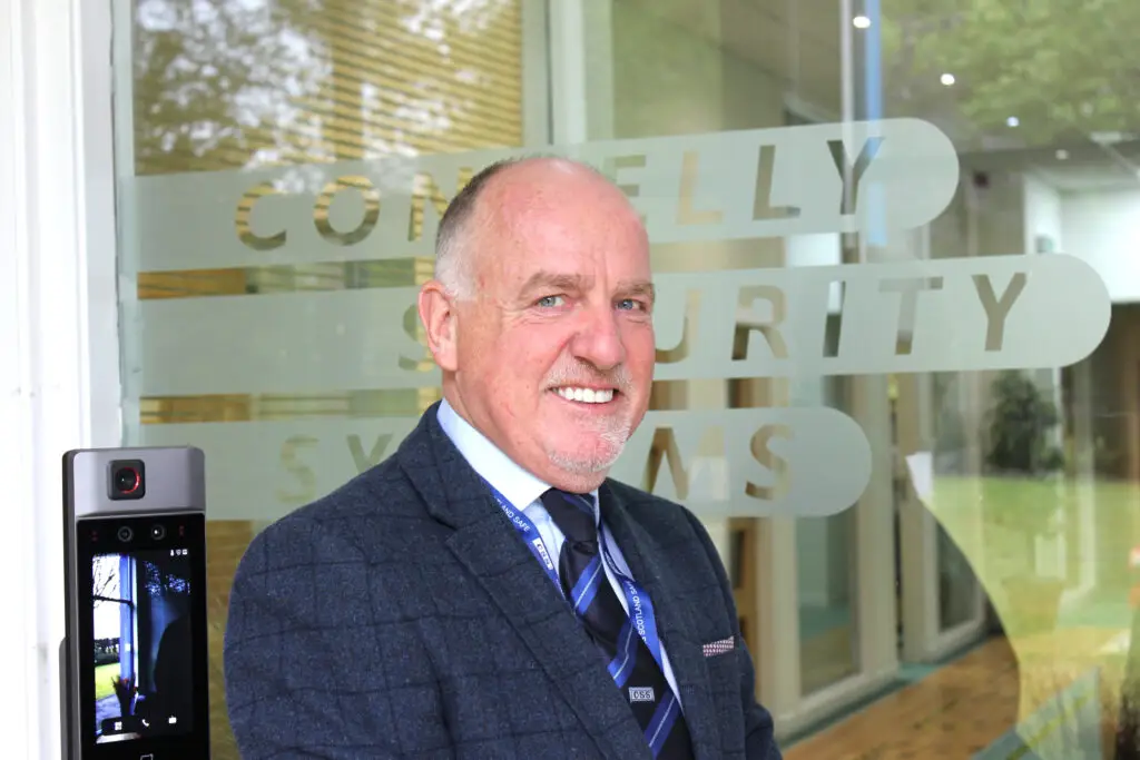 Photo of Paul Connelly smiling and standing in front of a glass door that says Connelly Security Systems. A door video entry screen is at the bottom left.