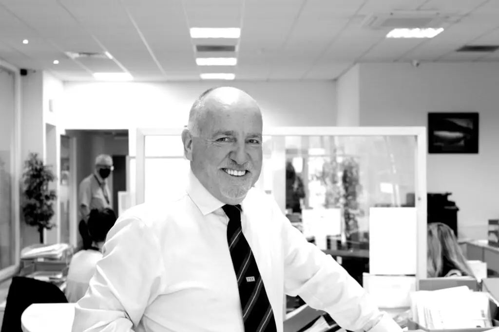 Black and white photo of Paul Connelly standing in CSS office resting his left hand against a desk. Members of staff working to social distance measures can be seen behind him, one of whom is wearing a black face mask. There are safety separator screens mounted to desks.