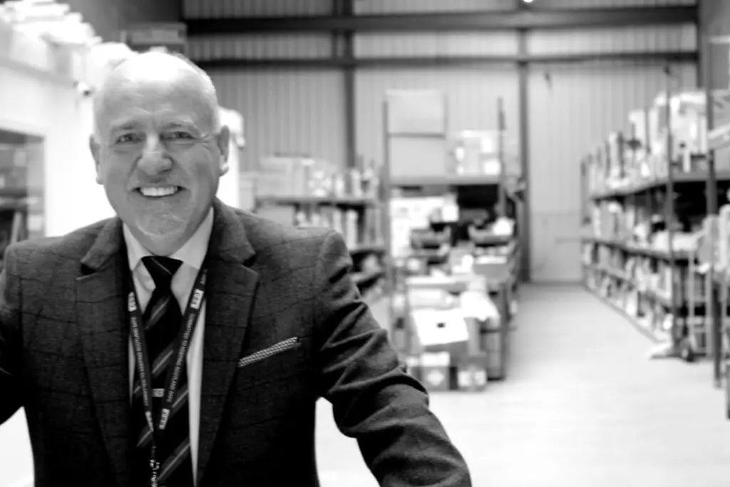 Black and white photo of Paul Connelly smiling and standing in a warehouse. Shelves, boxes and fire extinguishers are visible behind him.