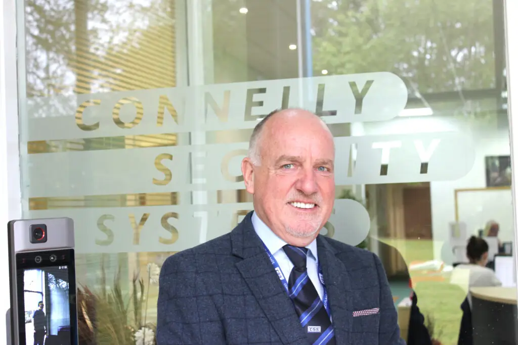 Photo of Paul Connelly smiling and standing in front of a glass door that says Connelly Security Systems. A door video entry screen is at the bottom left.