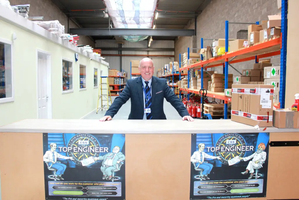 Photo of Paul Connelly smiling and standing at the counter in the warehouse. Shelves, boxes and fire extinguishers are visible behind him. Two posters on the counter front are designed like who wants to be a millionaire and text reads who wants to be a CSS top engineer.