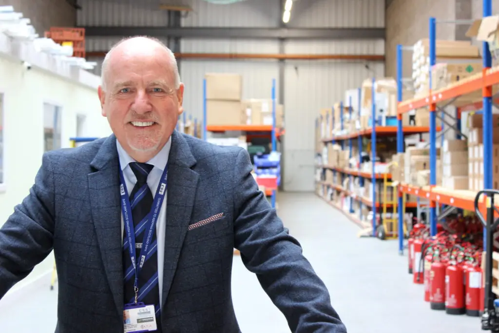 Photo of Paul Connelly smiling and standing in a warehouse. Shelves, boxes and fire extinguishers are visible behind him.