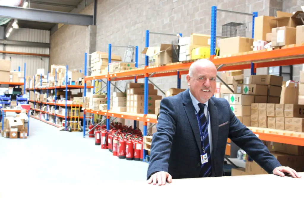 Photo of Paul Connelly smiling and standing at the counter in the warehouse. Shelves, boxes and fire extinguishers are visible behind him.
