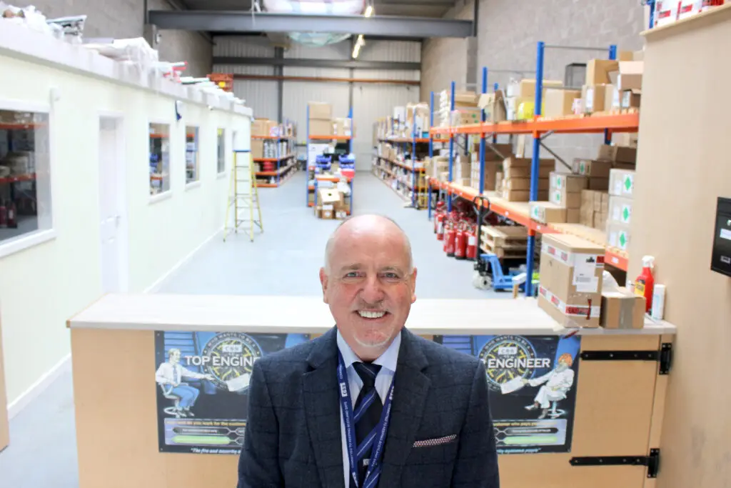 Photo of Paul Connelly smiling and standing in front of a counter in a warehouse. Shelves and boxes are visible behind him.