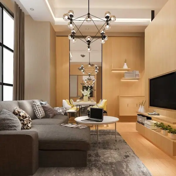 Side view of living room with grey L-shaped sofa with scatter cushions, white circular table with laptop, widescreen TV in wooden integrated wall unit with books and plants on shelves below, drop geometric lighting and dining table and chairs, wooden sideboard and wall mounted shelves in background.